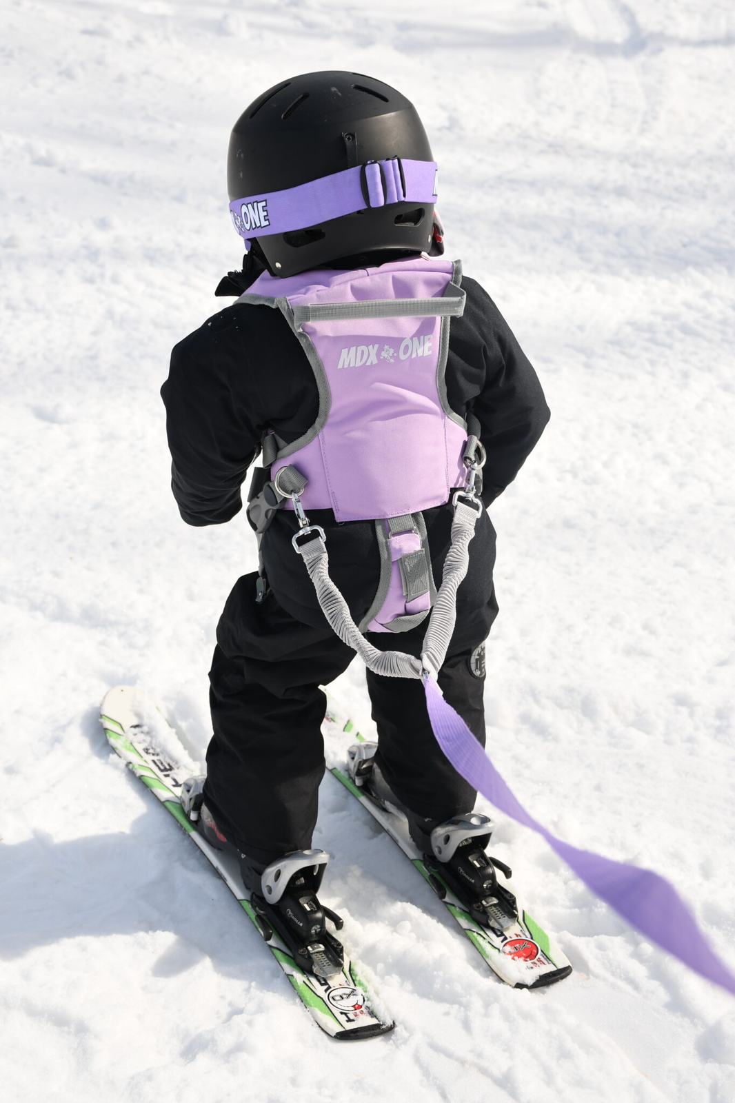 Bambino con attrezzatura da sci, casco nero, imbracatura viola e guinzaglio, in piedi sulla neve.